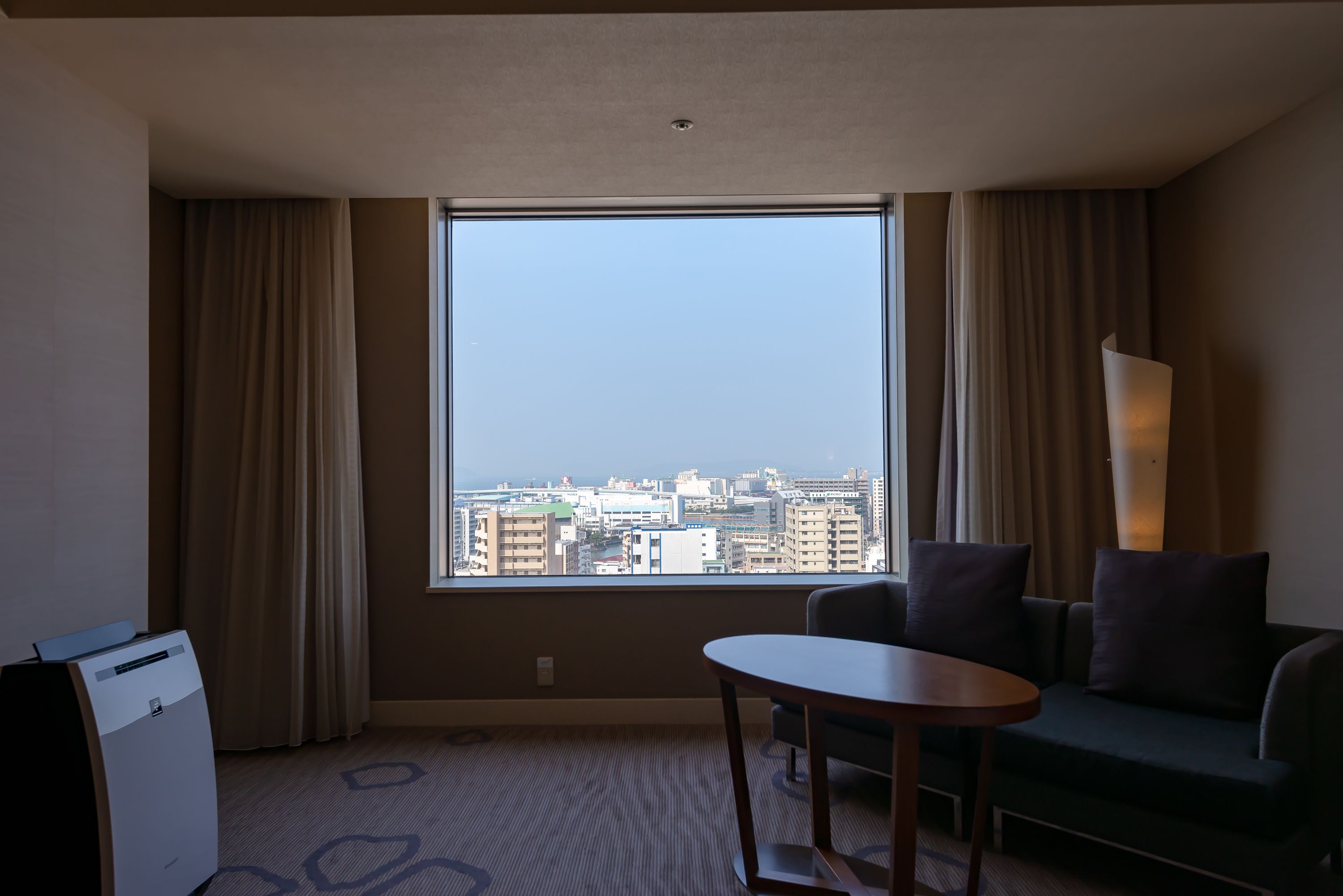 A hotel room with a large window offering a city view, a sofa, a table, and a lamp.