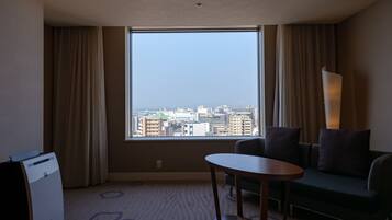 A hotel room with a large window offering a city view, a sofa, a table, and a lamp.