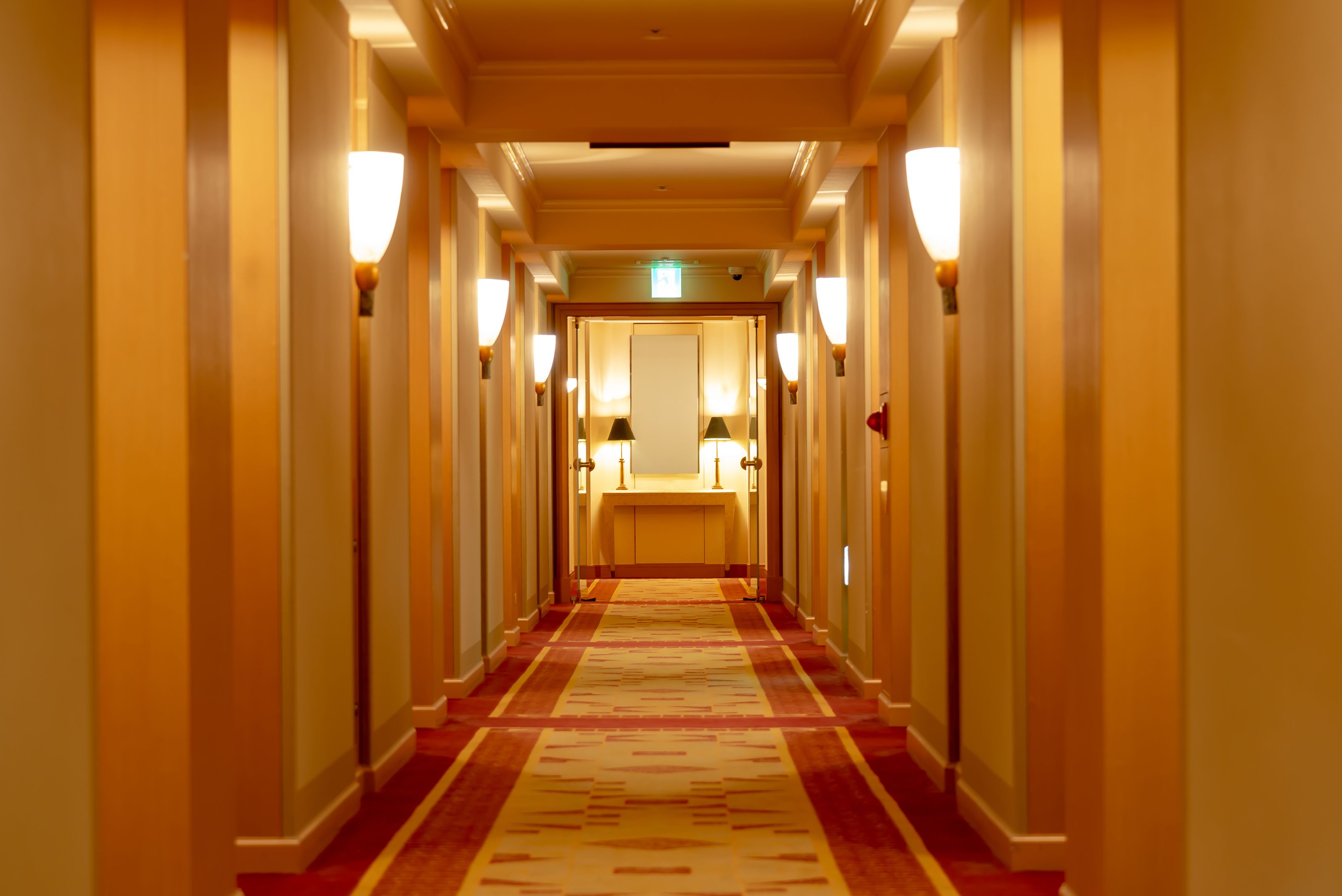 A hotel hallway with a carpeted floor, wall-mounted lamps, and a reception desk at the end.