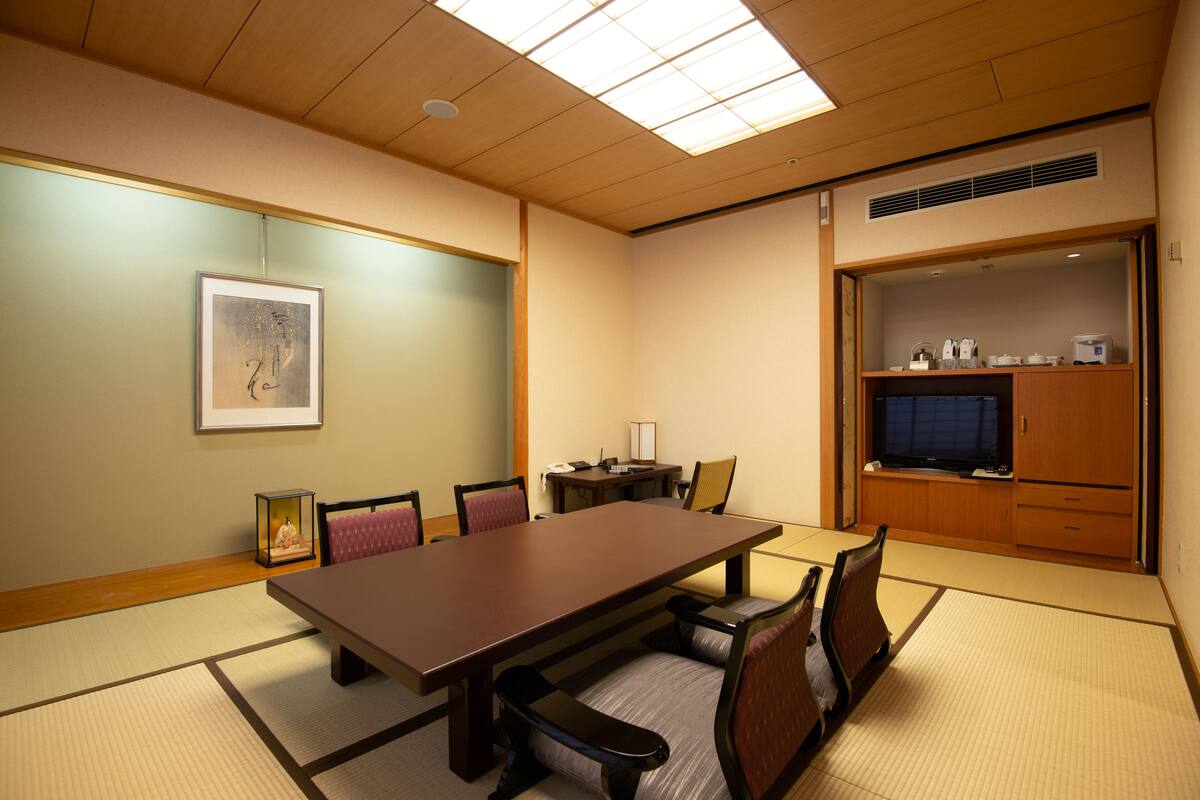 Une salle de conférence avec une longue table, des chaises, un tableau encadré au mur, un petit meuble contenant divers objets et un téléviseur.