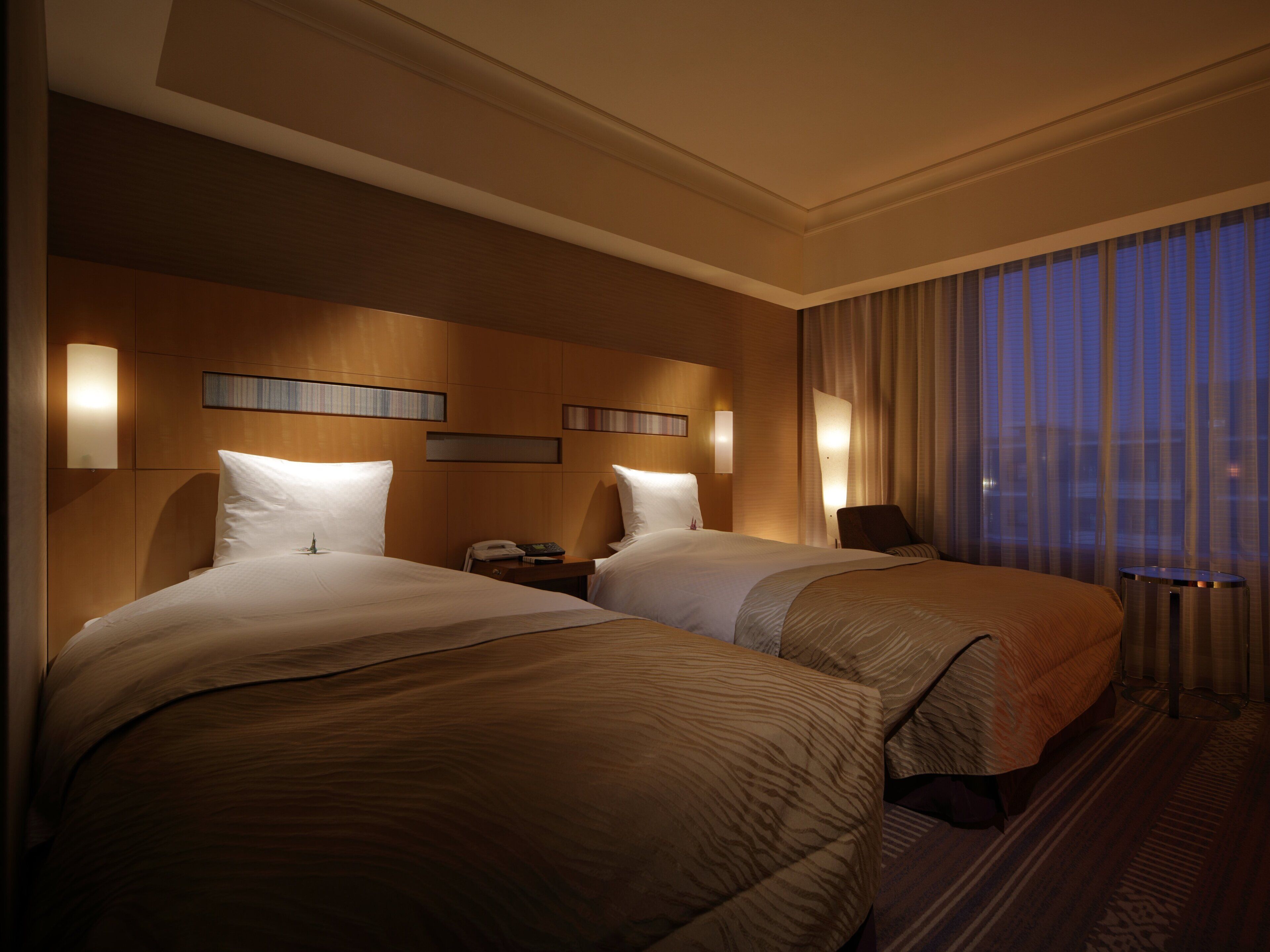 Une chambre d’hôtel avec deux lits, une table de chevet, une lampe et une vue sur la ville au crépuscule.