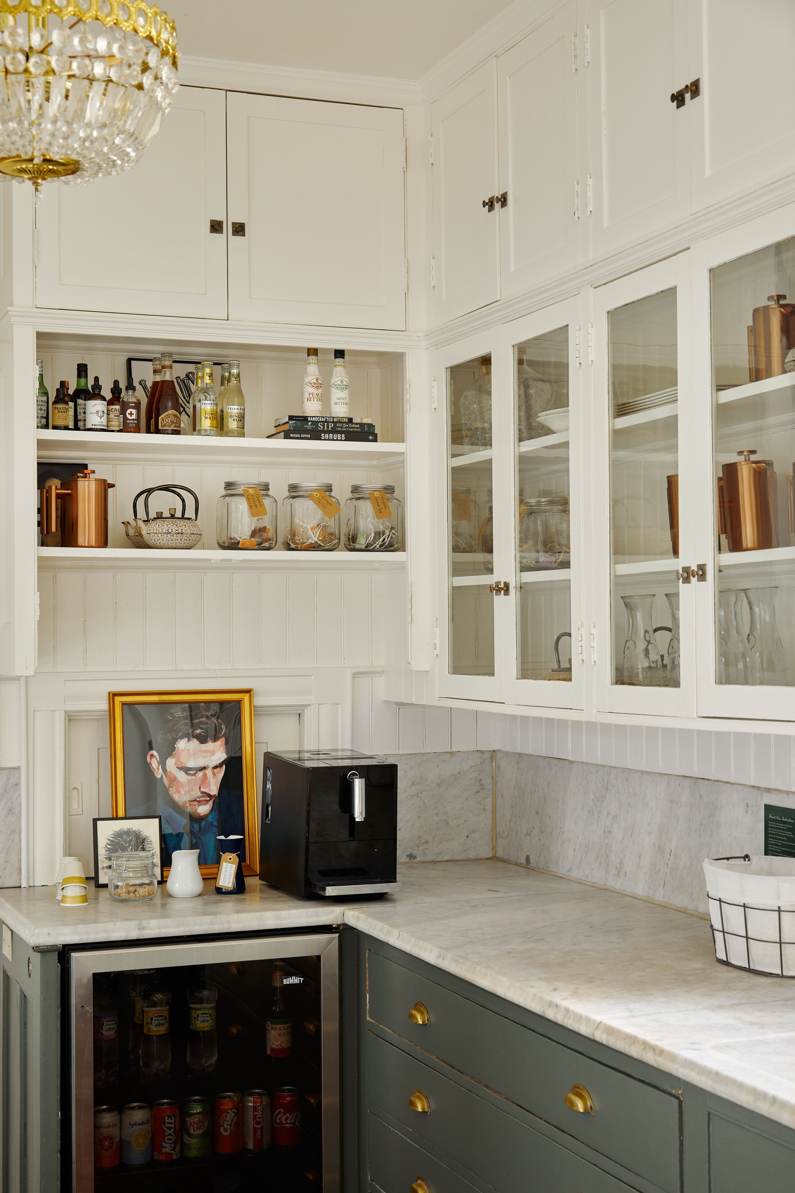 Eine Küche mit weißen Schränken, einem Kühlschrank und einer Mikrowelle. Regale mit Gläsern und Flaschen, und ein gerahmtes Bild auf der Arbeitsfläche.