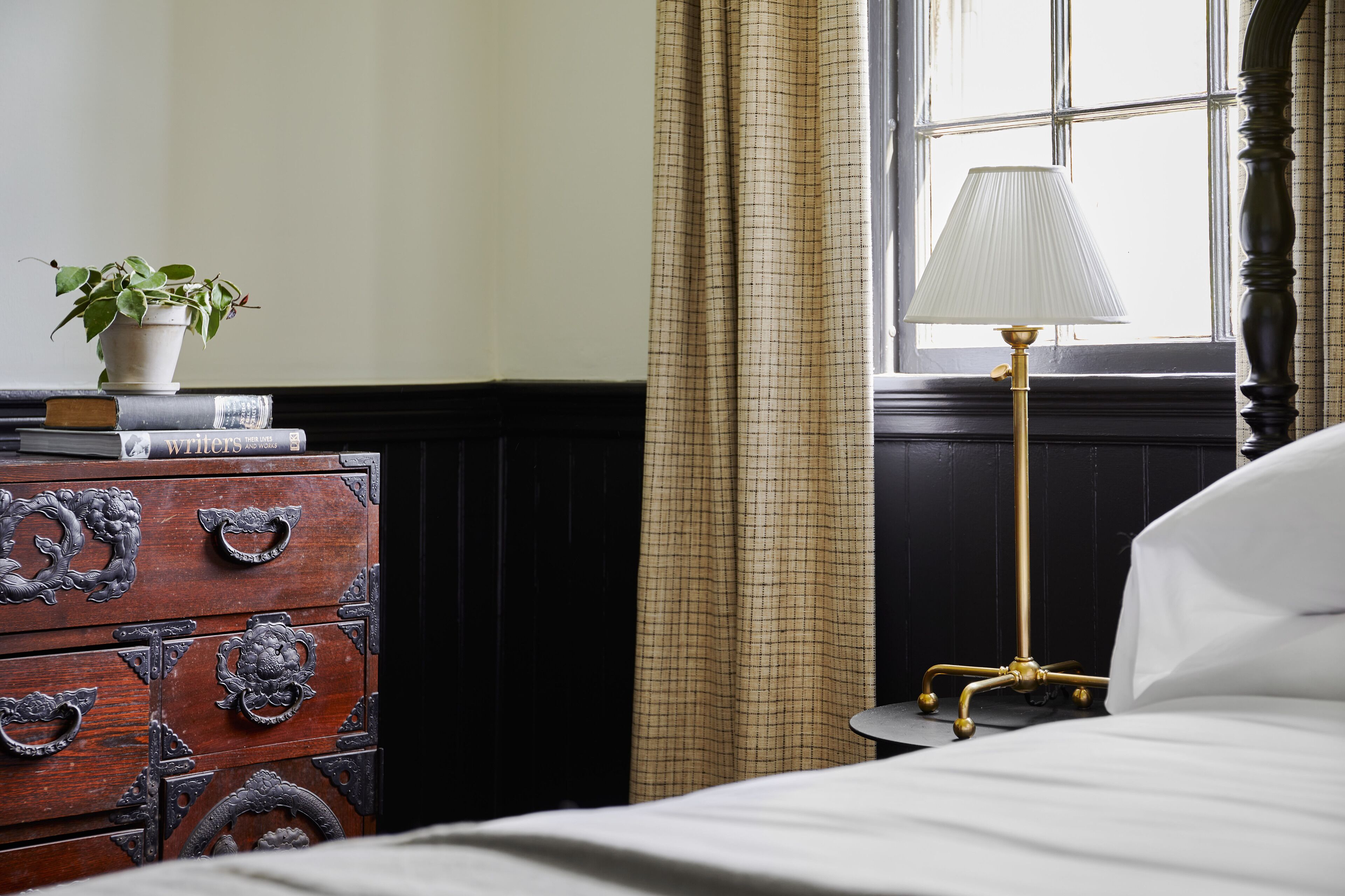 Una cama bien hecha con edredón blanco, una lámpara de noche, un aparador de madera con tiradores decorativos, una planta en maceta y una ventana con cortinas.