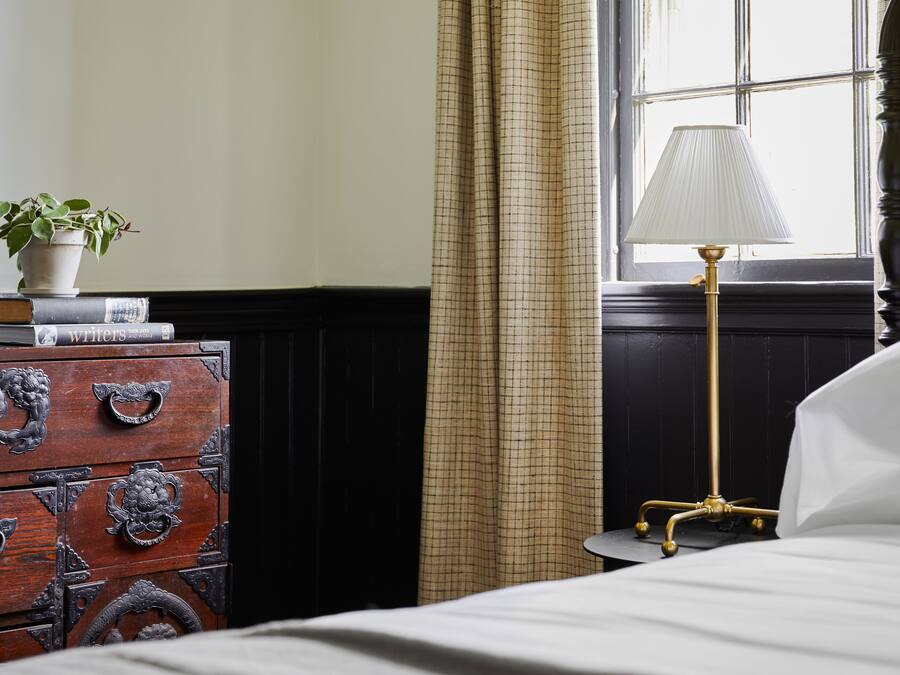 Una cama bien hecha con edredón blanco, una lámpara de noche, un aparador de madera con tiradores decorativos, una planta en maceta y una ventana con cortinas.