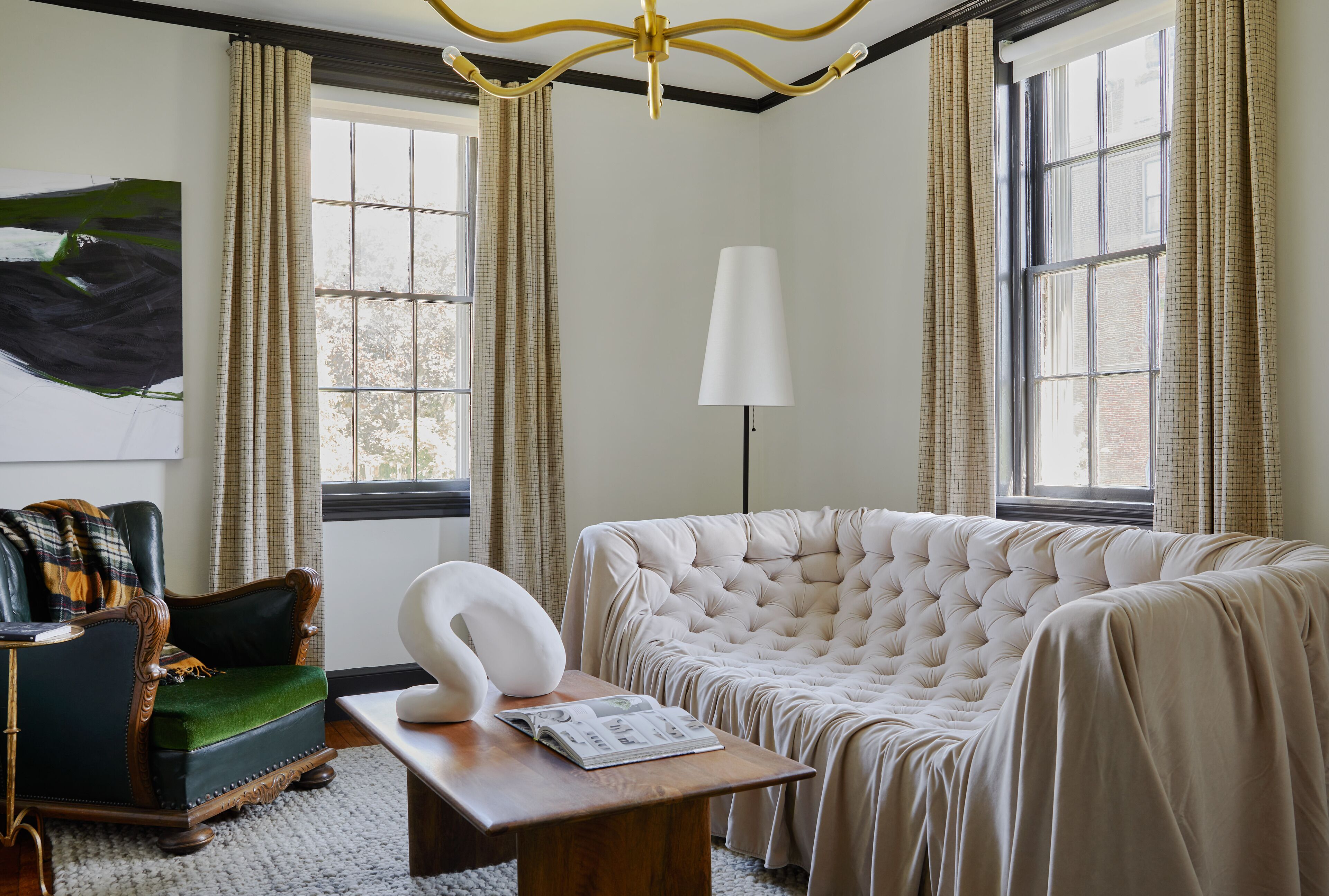 A room with a tufted sofa, a green armchair, a white lamp, and a large abstract painting on the wall.