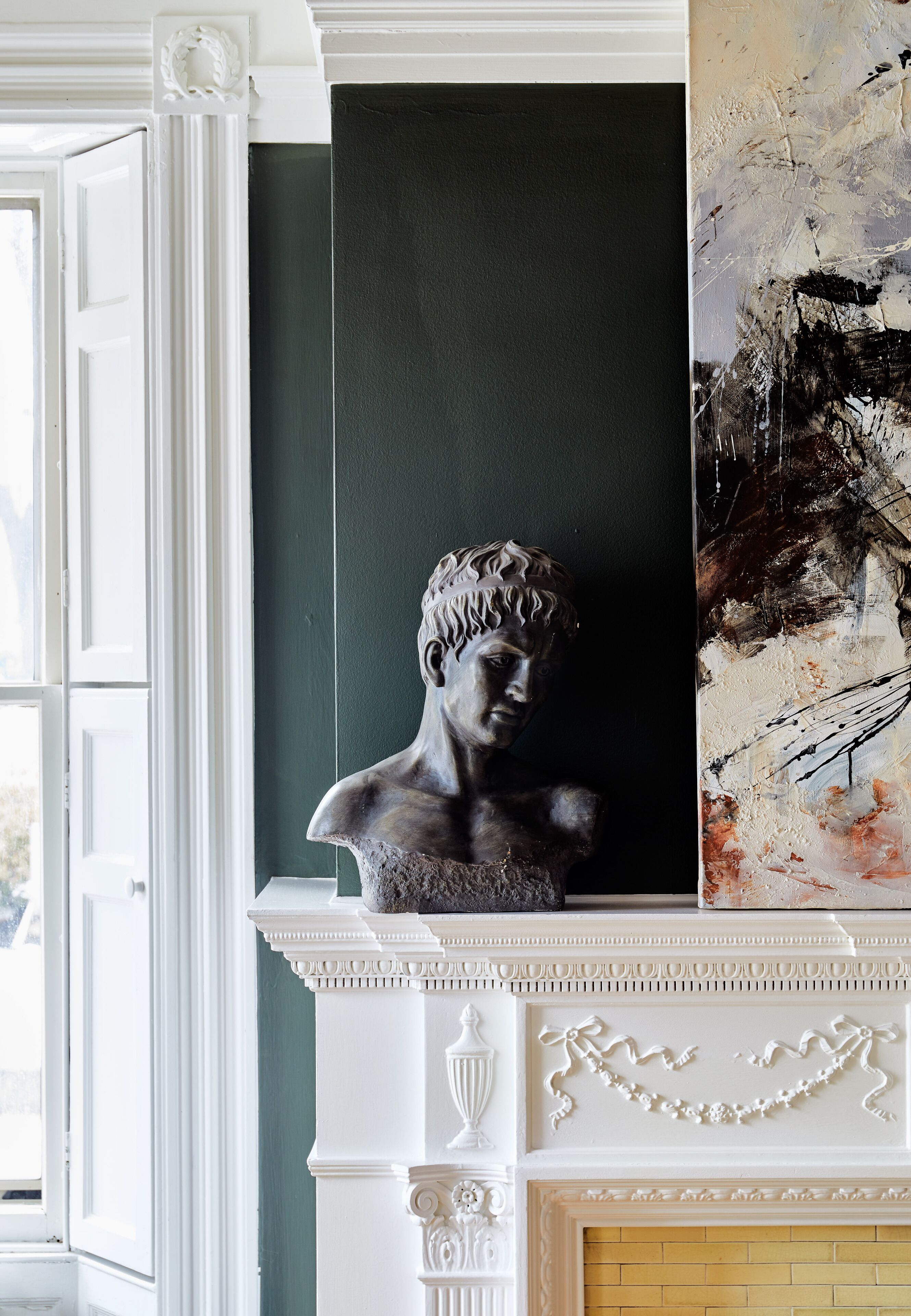 A room with a classical bust sculpture on a mantelpiece, a dark green wall, and a fireplace with decorative moldings.