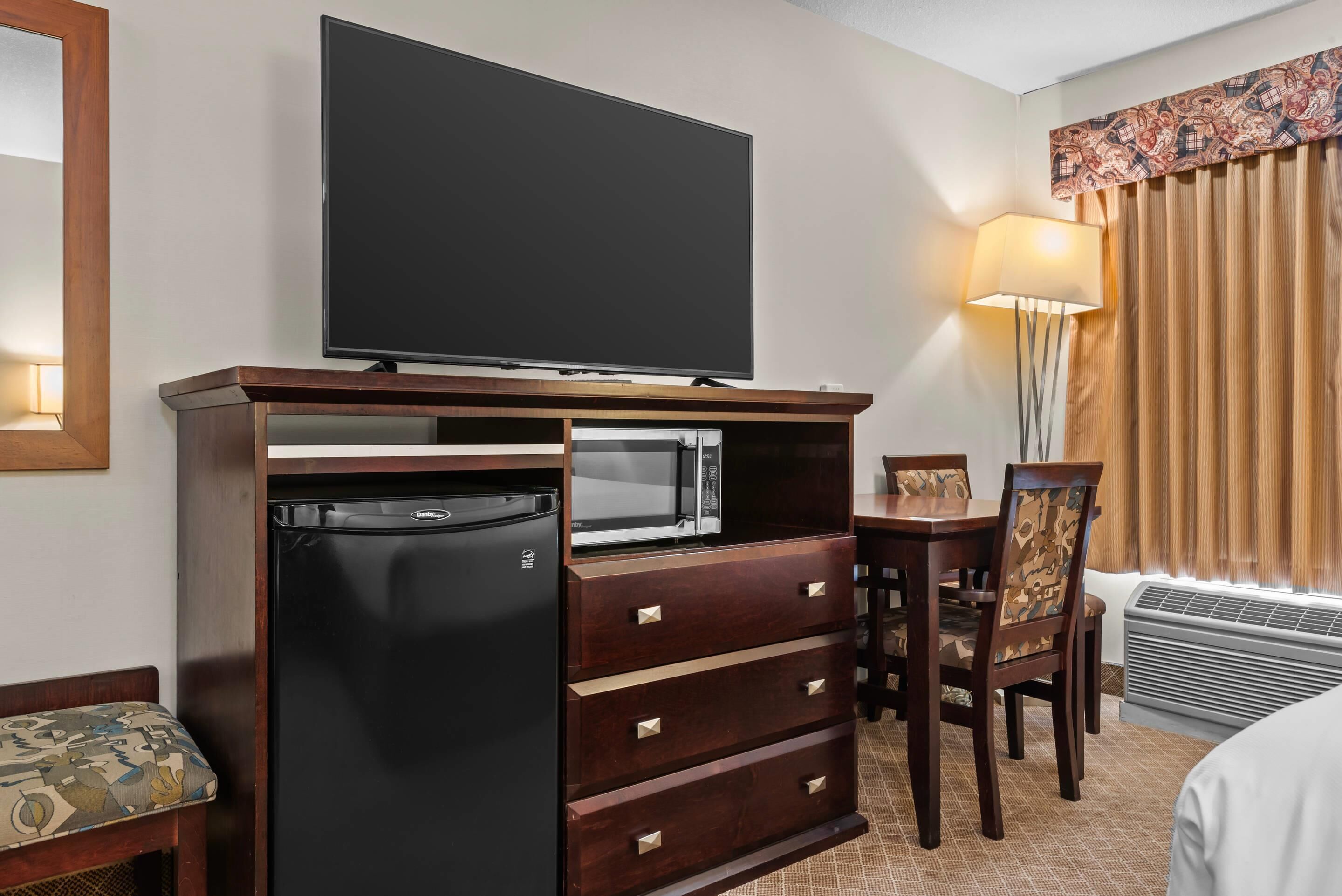 Hotelkamer met flatscreen televisie bevestigd boven een houten kast, een magnetron, een koelkast, een eettafel met stoelen, een lamp en een raam met gordijnen.