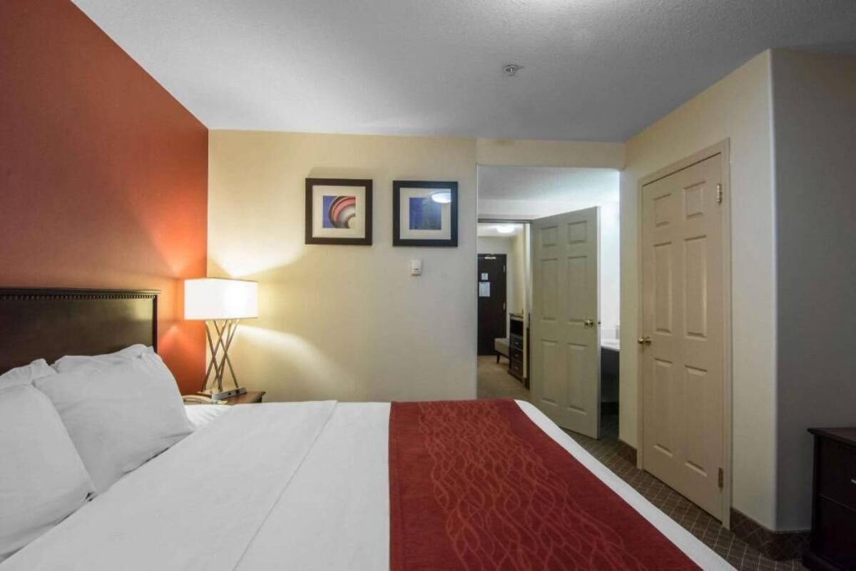 A hotel room with a bed, a nightstand, a lamp, two framed pictures on the wall, and a door leading to another room.