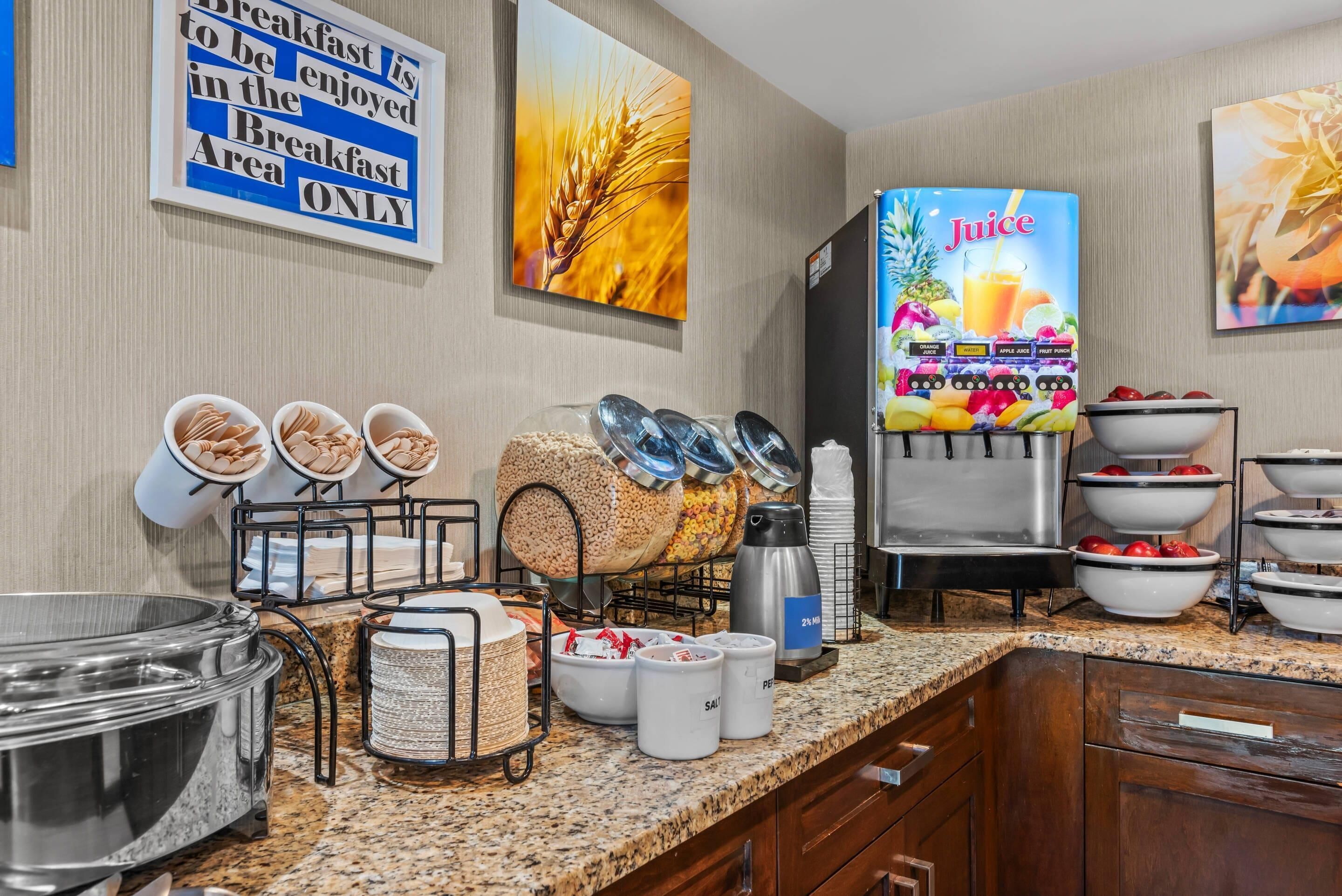 A breakfast buffet area with cereal bowls, a juice dispenser, and a sign indicating breakfast is served in the area only.