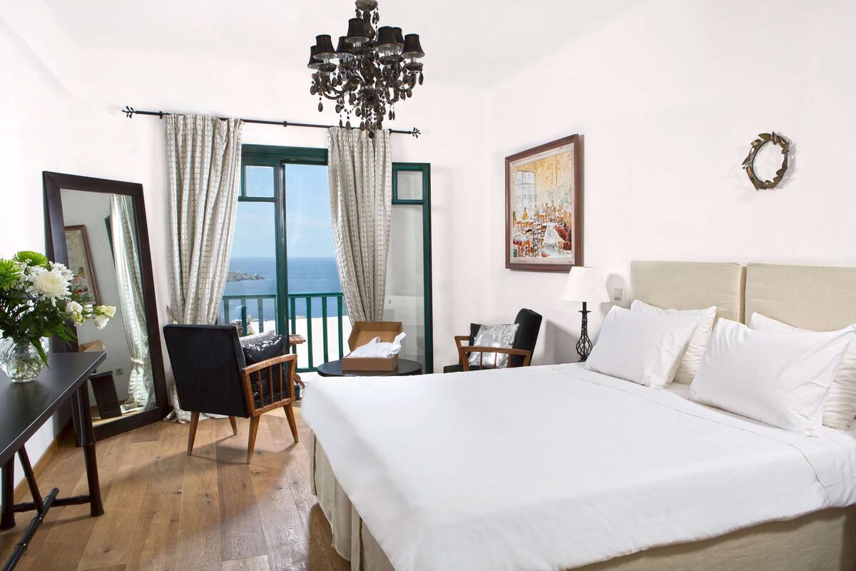 A bedroom with a large bed, a desk, a chair, a mirror, and a view of the sea.