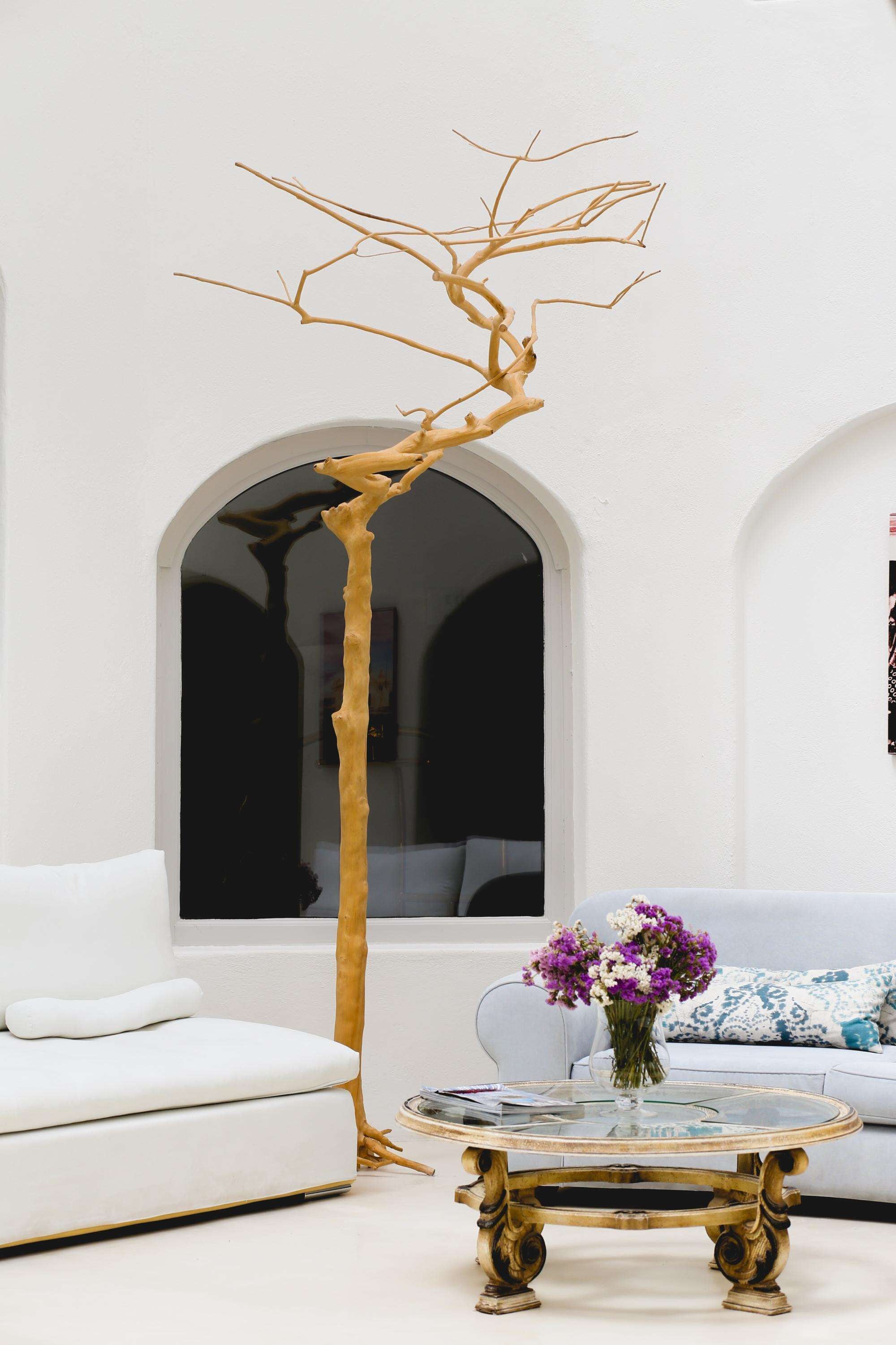 A modern living room with a white sofa, a glass coffee table with gold legs, and a unique tree sculpture.