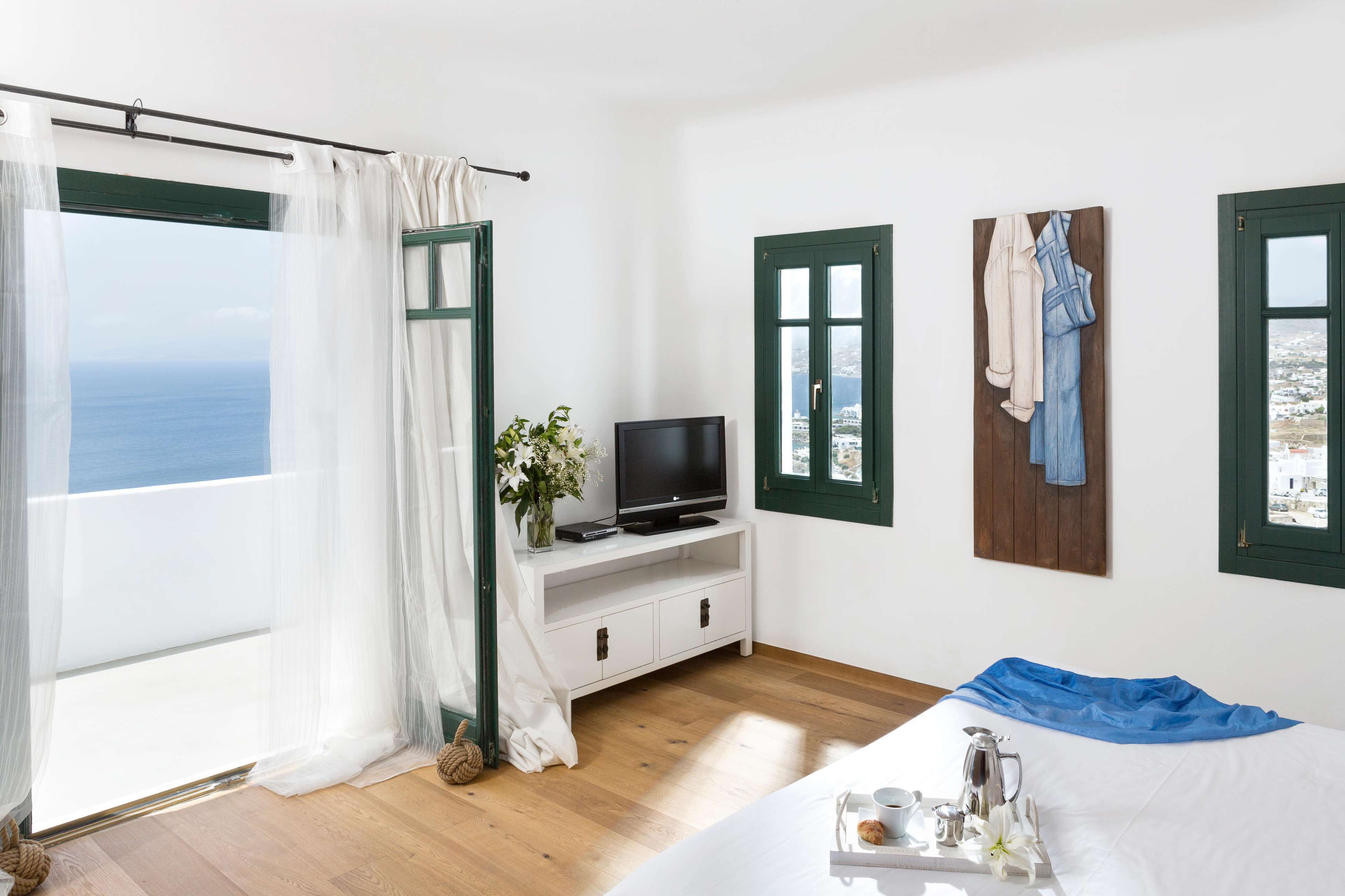 Una habitación con un ventanal grande, un televisor sobre un mueble blanco, una cama con una toalla azul y un perchero en la pared.