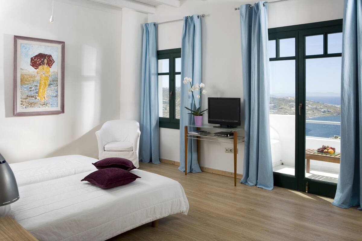 A bedroom with a bed, a desk with a computer, a chair, and a view of the sea.