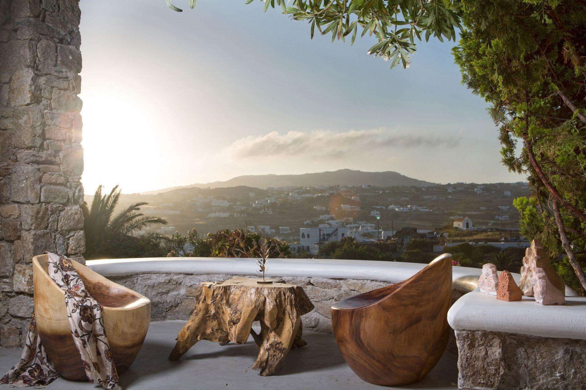 A stone table with a rustic wooden chair and a small wooden stool in a scenic outdoor setting with a view of a town and hills in the background.