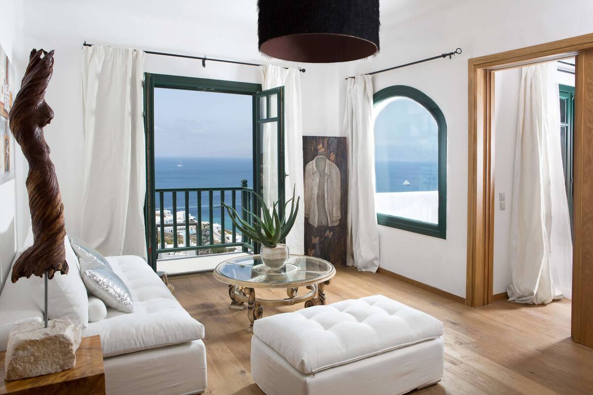 A room with a white sofa, a round glass table, and a view of the ocean through an open door.