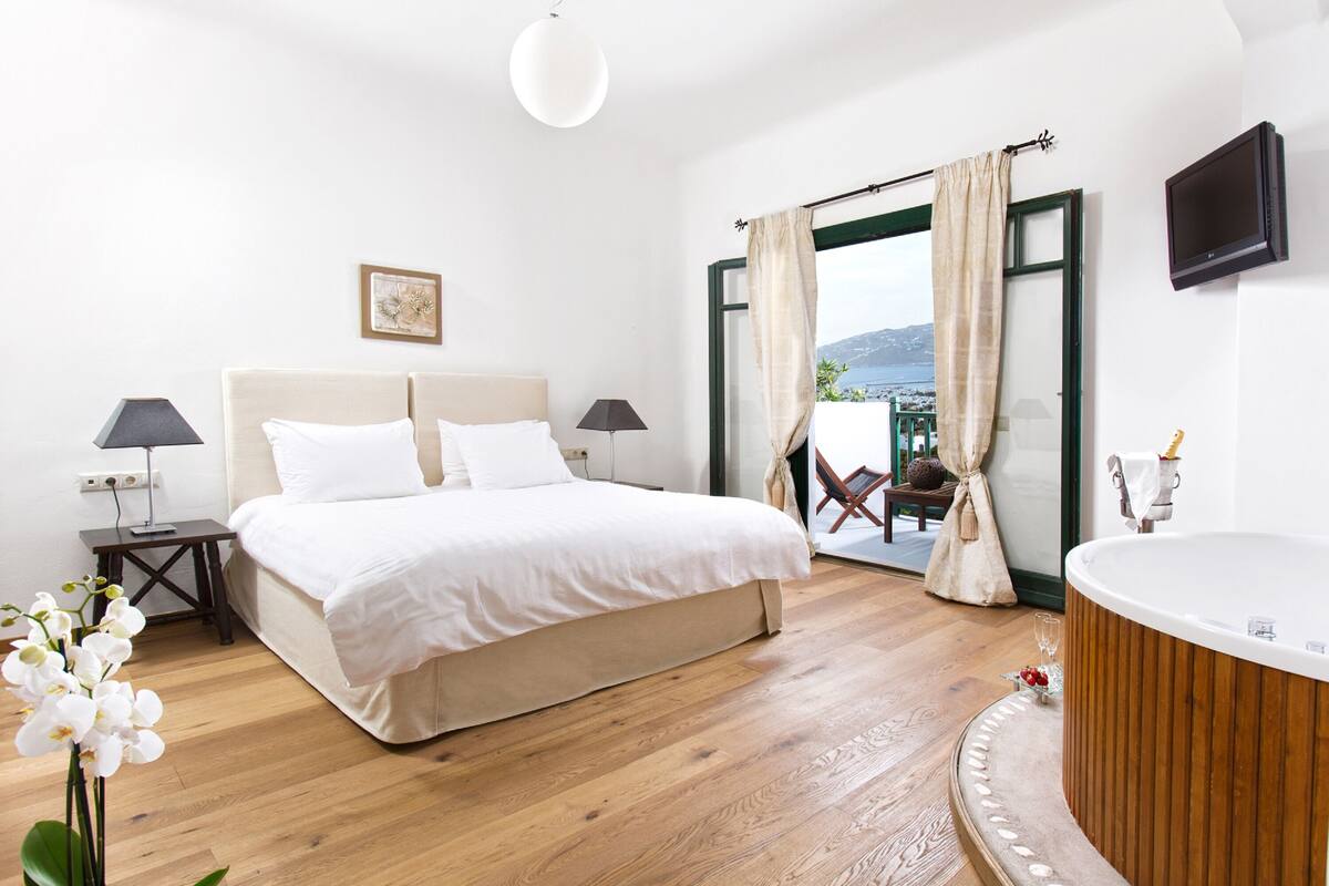 A bedroom with a bed, bedside lamps, a TV, a bathtub, and a view of the outdoors.