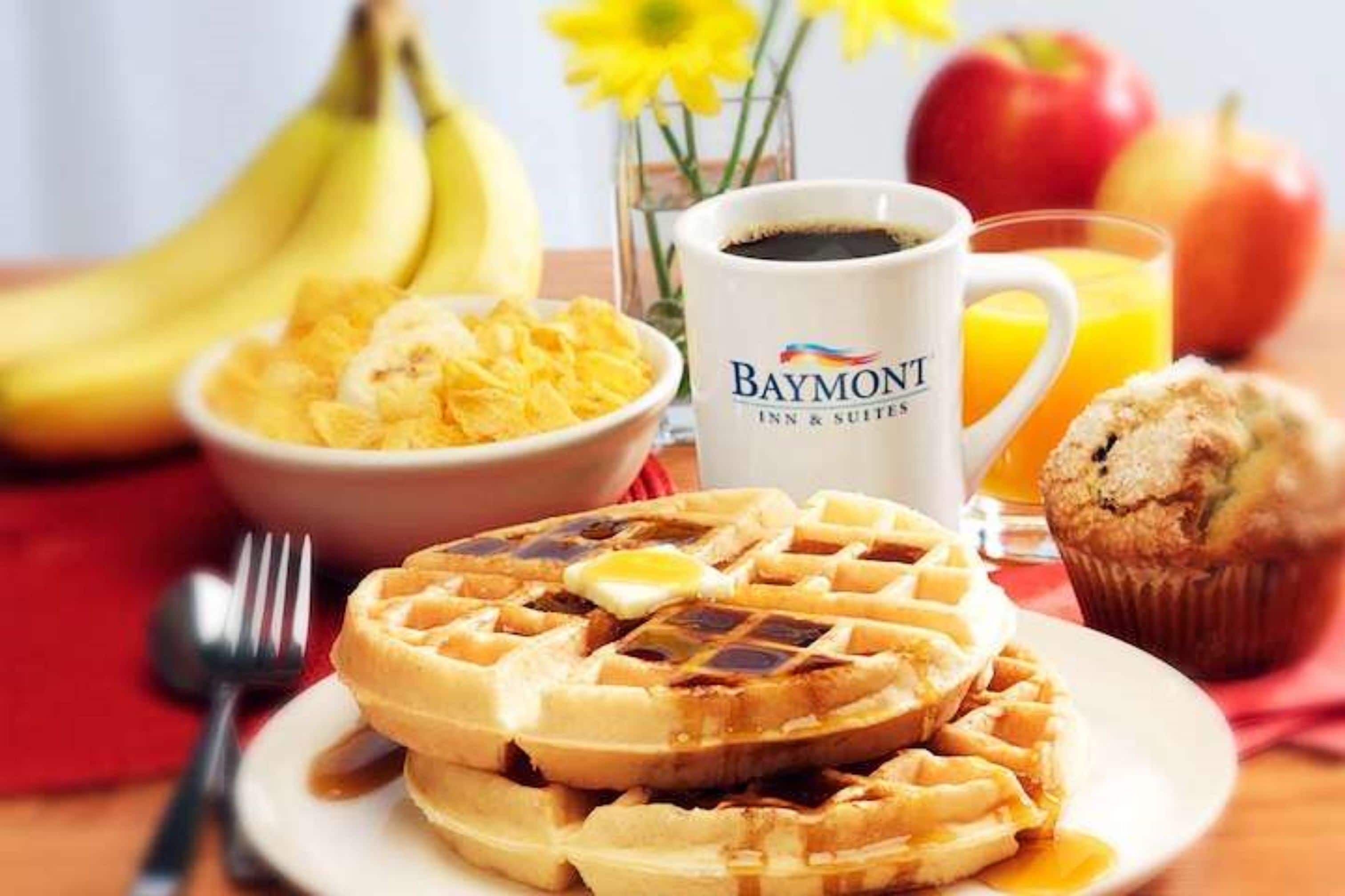 A breakfast setting with waffles, a muffin, a bowl of cereal, a cup of coffee, bananas, apples, and a glass of orange juice.