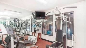 Un gimnasio con diversas máquinas de ejercicio, un televisor y grandes ventanales.