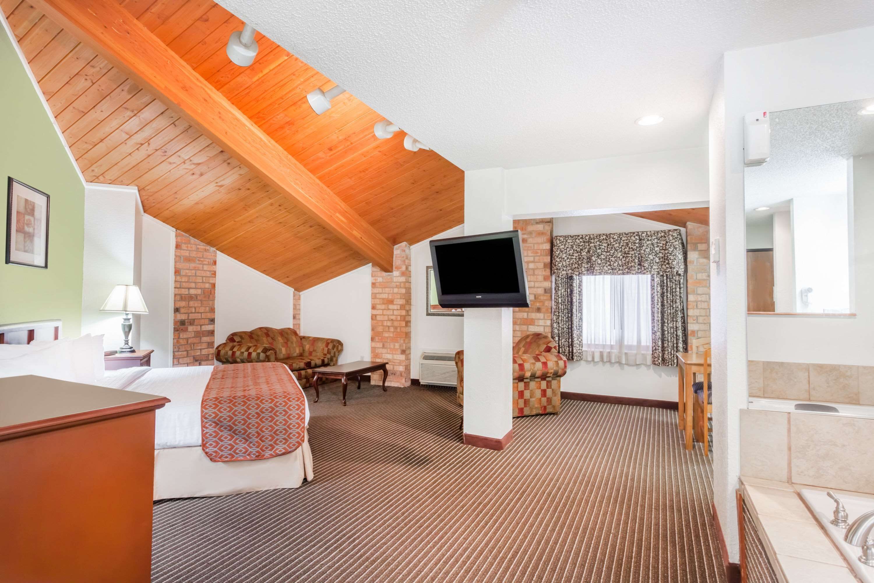 A hotel room with a sloped ceiling, a flat-screen TV, a sofa, a chair, a small table, and a bathroom with a sink and bathtub.
