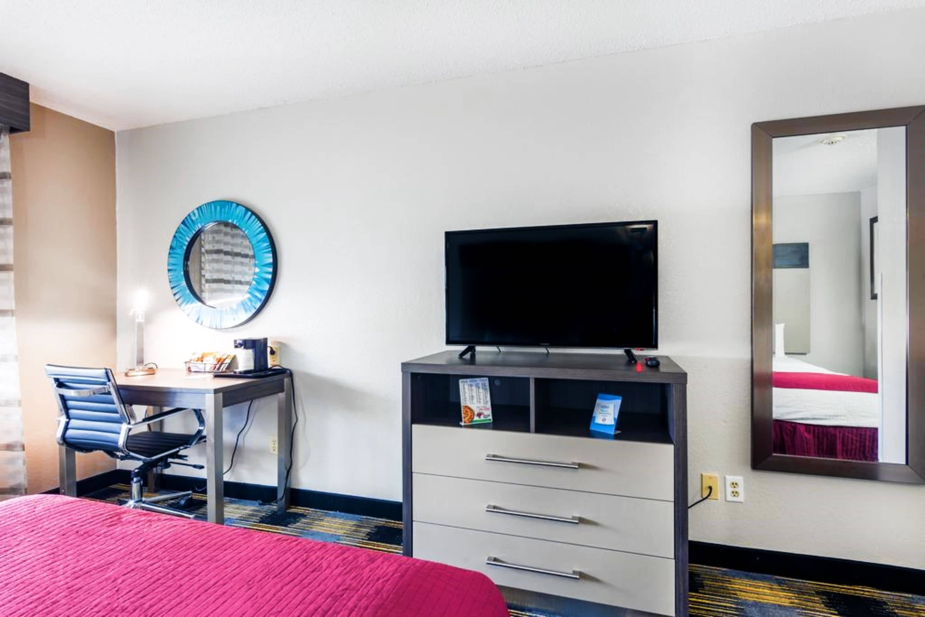 A hotel room with a bed, a desk with a chair, a television, a dresser, a mirror, and a window.