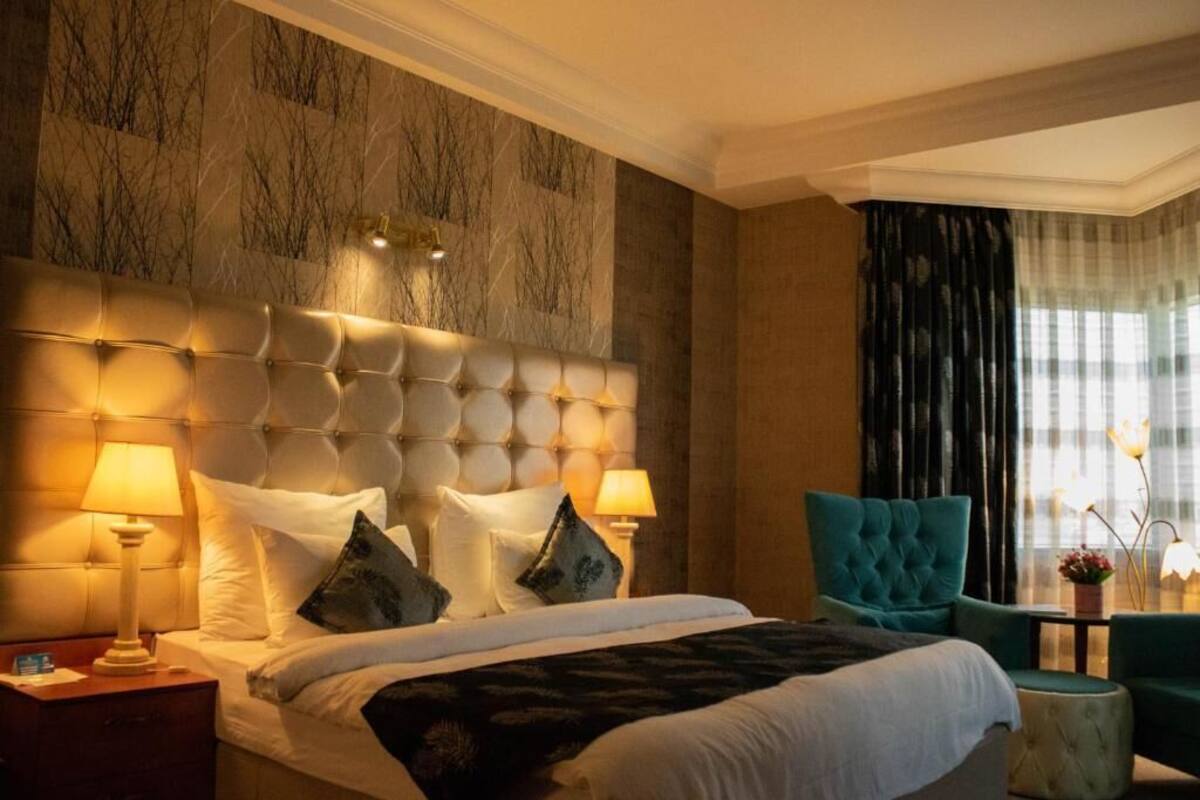 A hotel room with a tufted headboard, two bedside lamps, a bed with white linens, a teal armchair, and a view of the outdoors through sheer curtains.