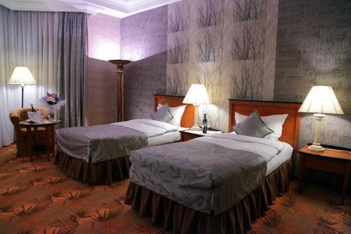 A hotel room with two beds, a large window with curtains, a round table with a lamp, and a wall with a textured pattern.