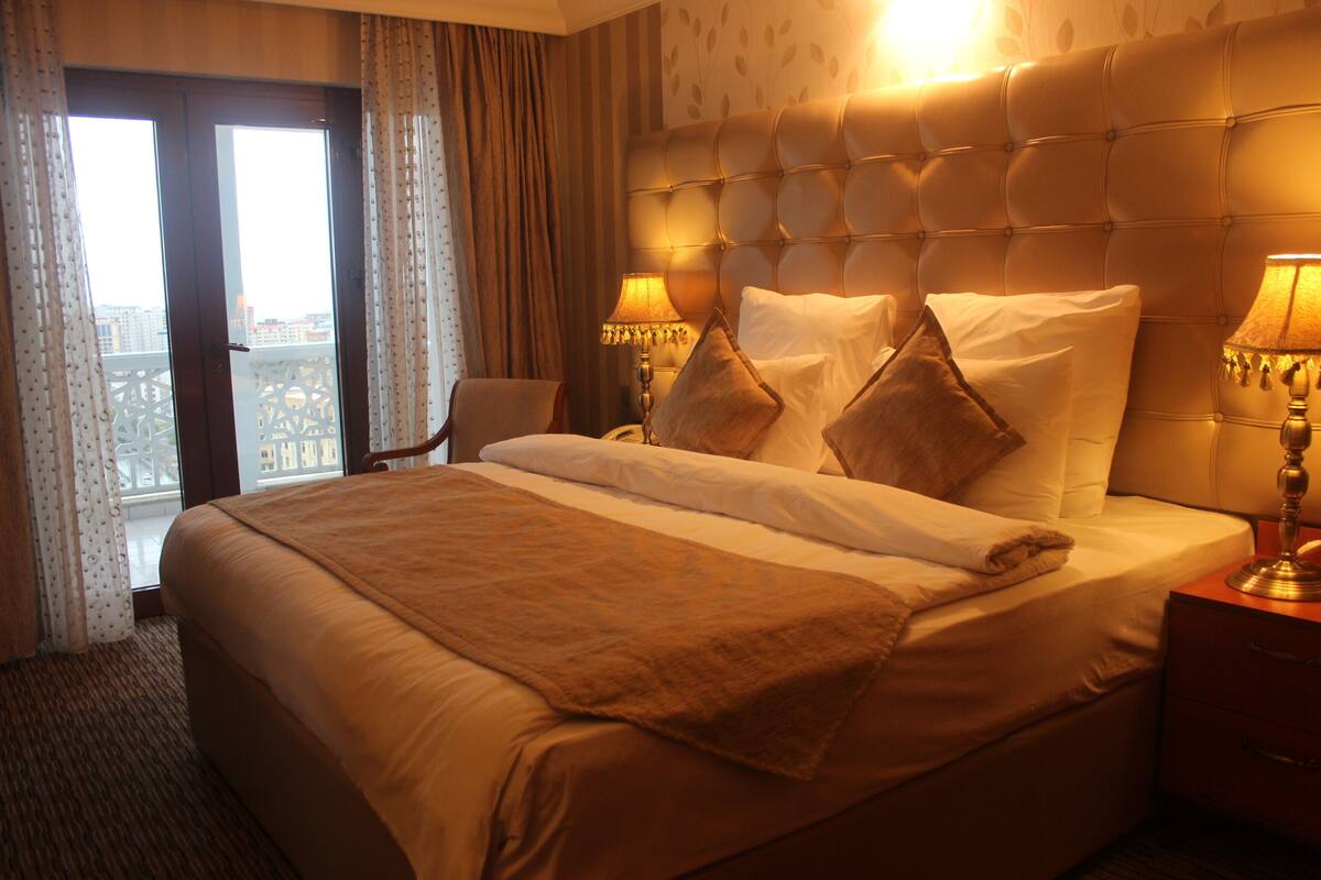 A hotel room with a large bed, two bedside lamps, a wooden nightstand, and a view of the city through the window.