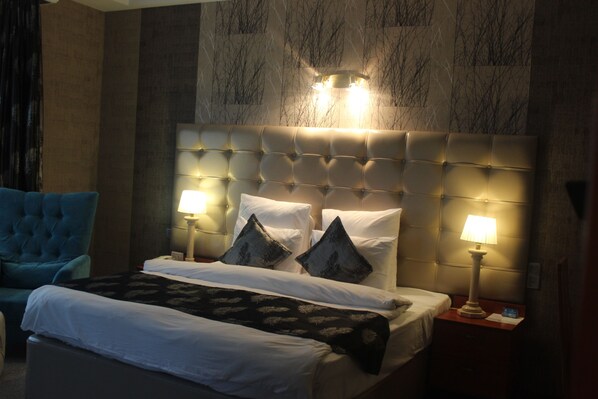 A hotel room with a tufted headboard, two bedside lamps, a bed with white linens, a blue armchair, and a patterned throw blanket. - Grand Hotel Europe (Baku)