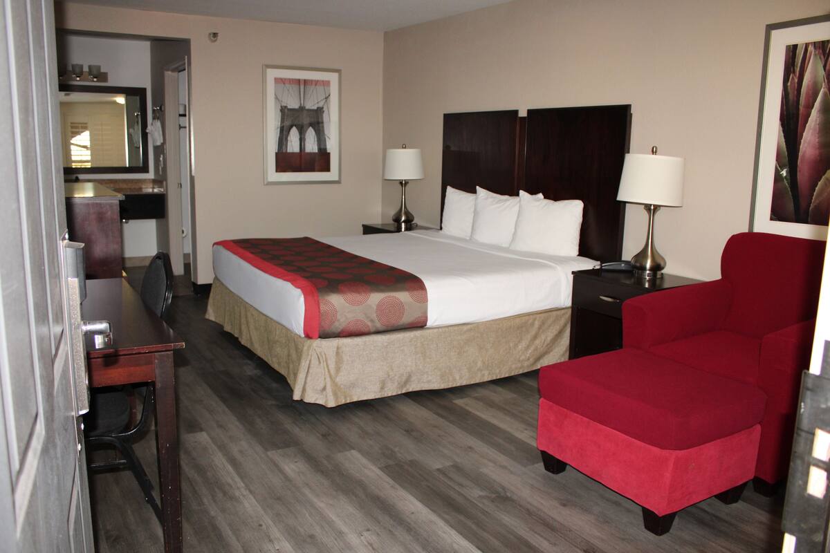 A hotel room with a bed, two bedside tables with lamps, a red armchair, a desk with a chair, a mirror, and a framed picture on the wall.