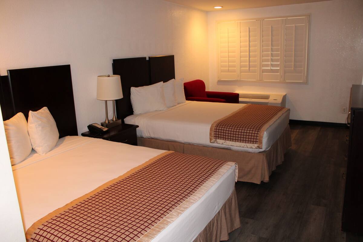 A hotel room with two beds, a nightstand with a lamp, a red chair, and a window with blinds.