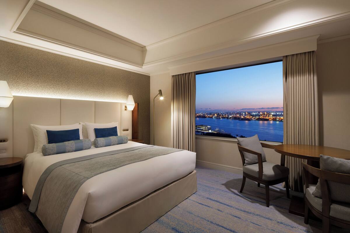A hotel room with a large bed, a desk, and a view of a cityscape and water.