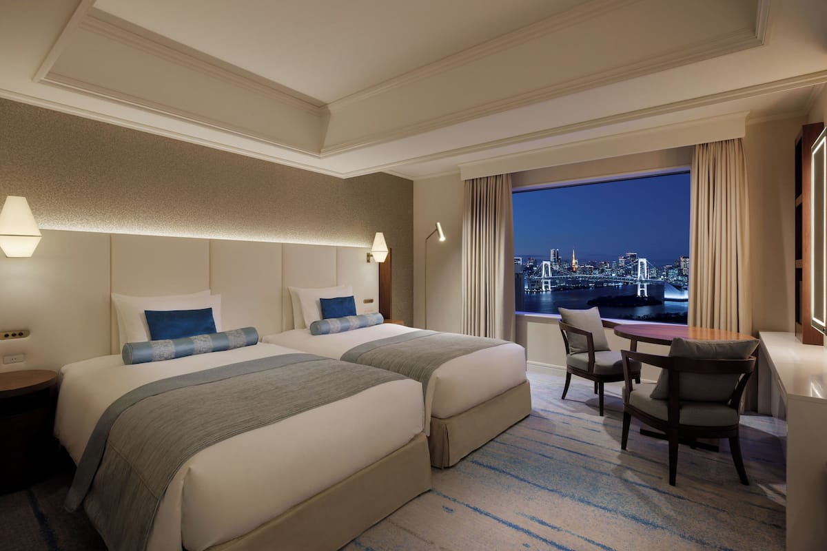 A hotel room with two beds, a desk, chairs, and a view of a cityscape at night.