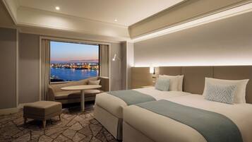 A hotel room with a large bed, a small sofa, a desk, and a view of a cityscape at dusk.