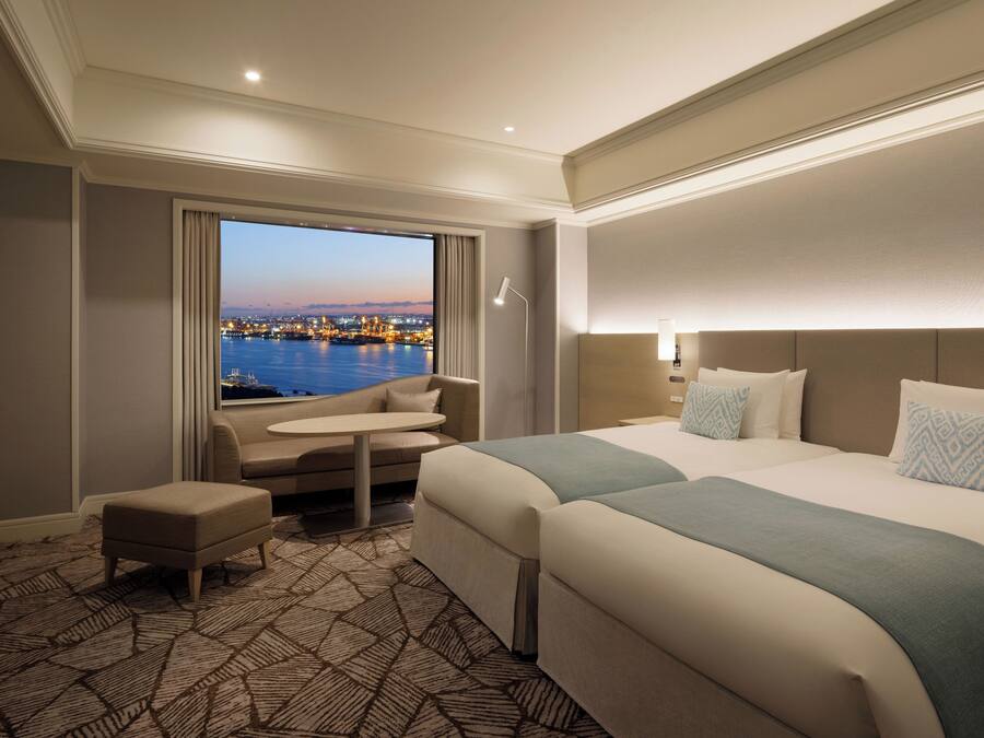 A hotel room with a large bed, a small sofa, a desk, and a view of a cityscape at dusk.