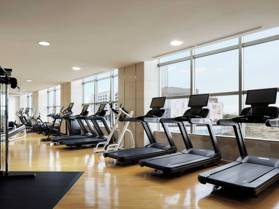 A gym with multiple treadmills and exercise bikes, large windows, and a wooden floor.