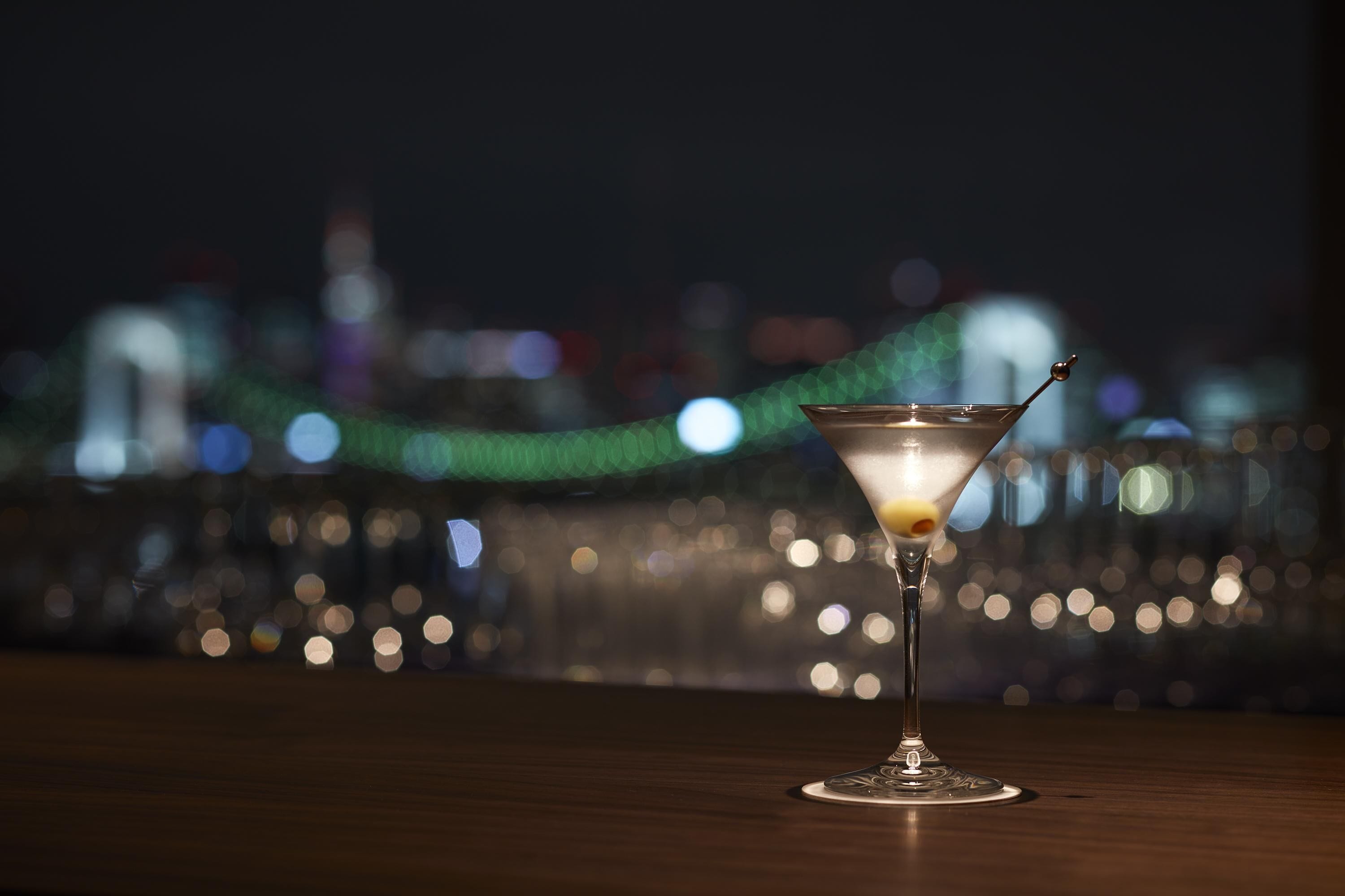 A martini glass on a wooden surface with a blurred cityscape in the background.