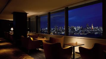 Un salón tenuemente iluminado con sillones de cuero y una mesa, grandes ventanales que ofrecen vistas nocturnas de la ciudad.