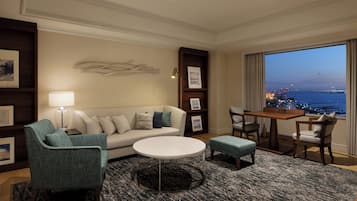 Une chambre d’hôtel avec un canapé, des fauteuils, une petite table, une table basse ronde, une bibliothèque et une vue sur la ville au crépuscule.
