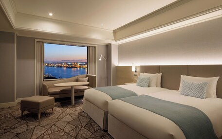 A hotel room with a large bed, a small sofa, a table, and a view of a cityscape at dusk.