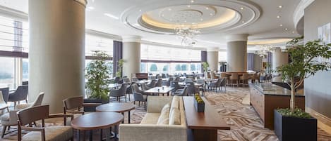 A spacious hotel lobby with high ceilings, large windows, and a central seating area with tables and chairs.
