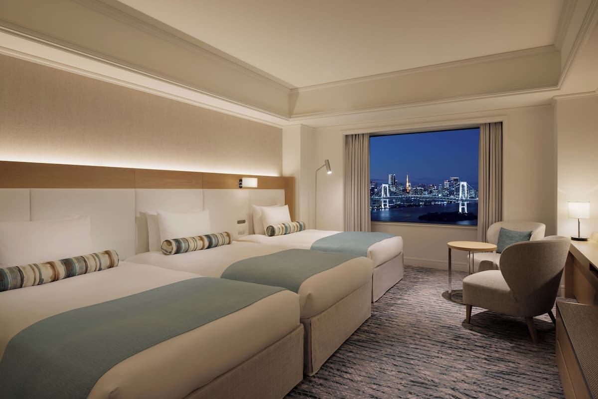 A hotel room with two beds, a chair, a small table, and a view of a cityscape at night.