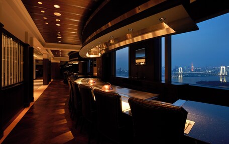 A restaurant with a view of a city skyline, featuring a long counter with chairs and a high ceiling with recessed lighting.