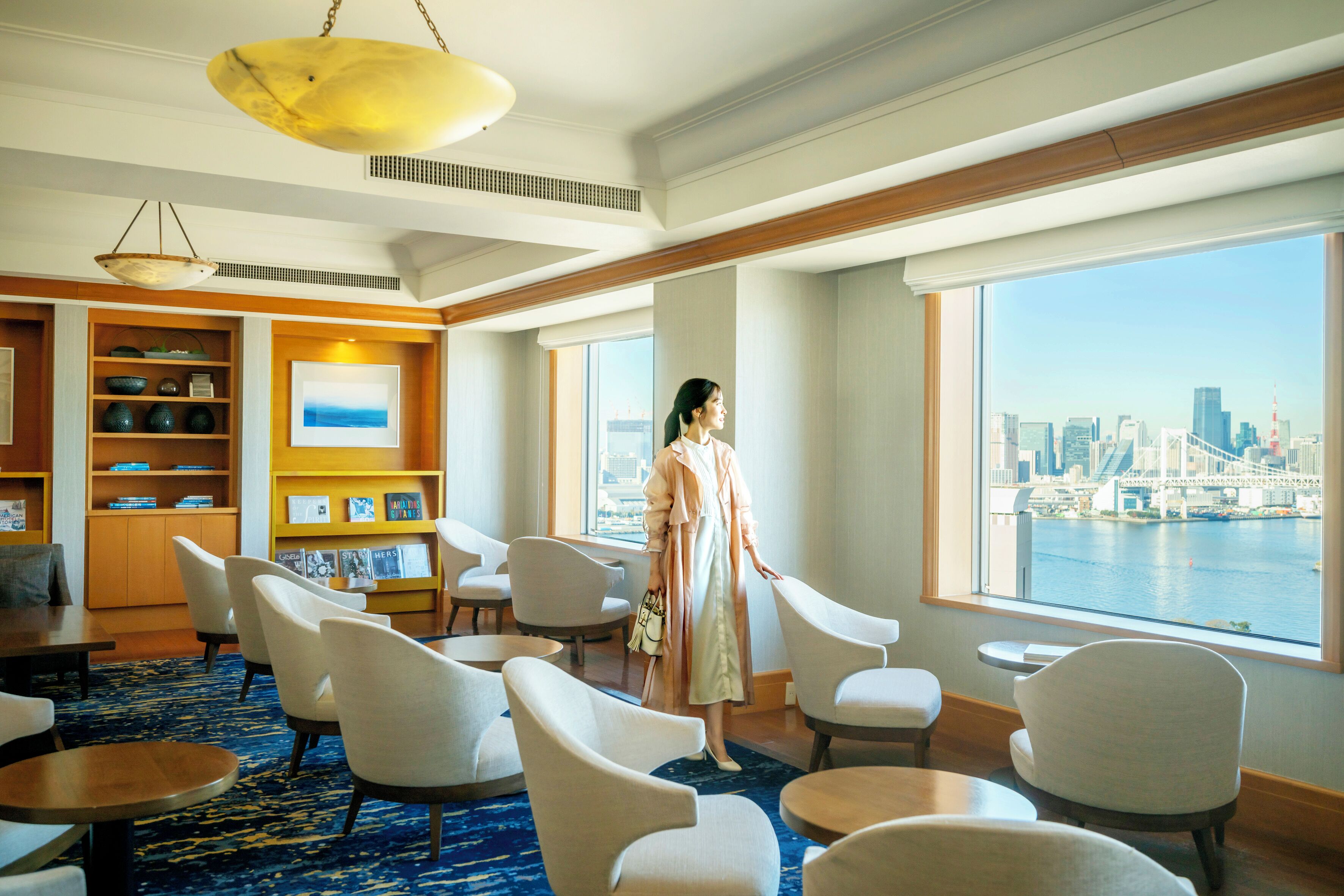 A hotel lobby with a large window offering a city view, a seating area with chairs and tables, and a bookshelf with books and decorative items.