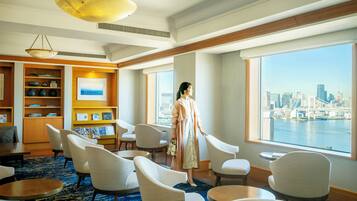 A hotel lobby with a large window offering a city view, a seating area with chairs and tables, and a bookshelf with books and decorative items.