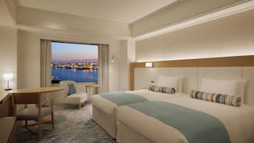 A hotel room with a large bed, a desk, a chair, and a view of a cityscape at dusk.