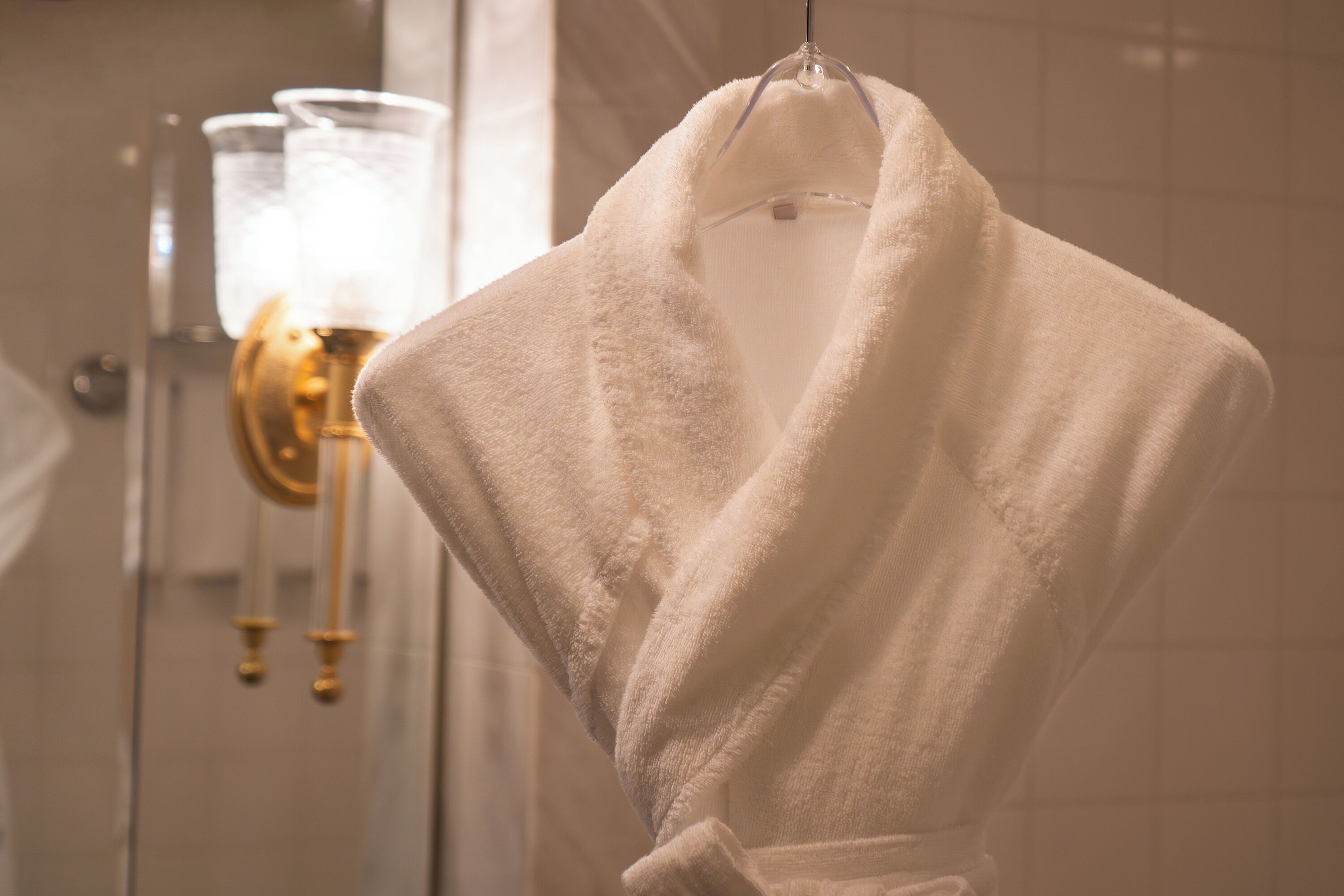 A white bathrobe hanging on a hook in a bathroom with a gold faucet and a glass light fixture.
