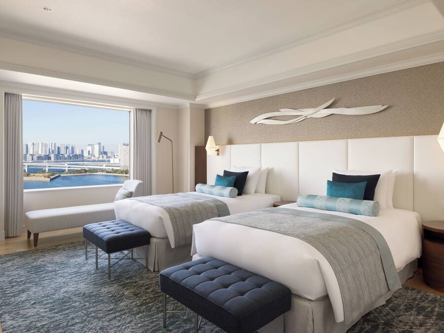 A hotel room with two beds, a bench, a view of a city skyline, and a cityscape visible through the window.