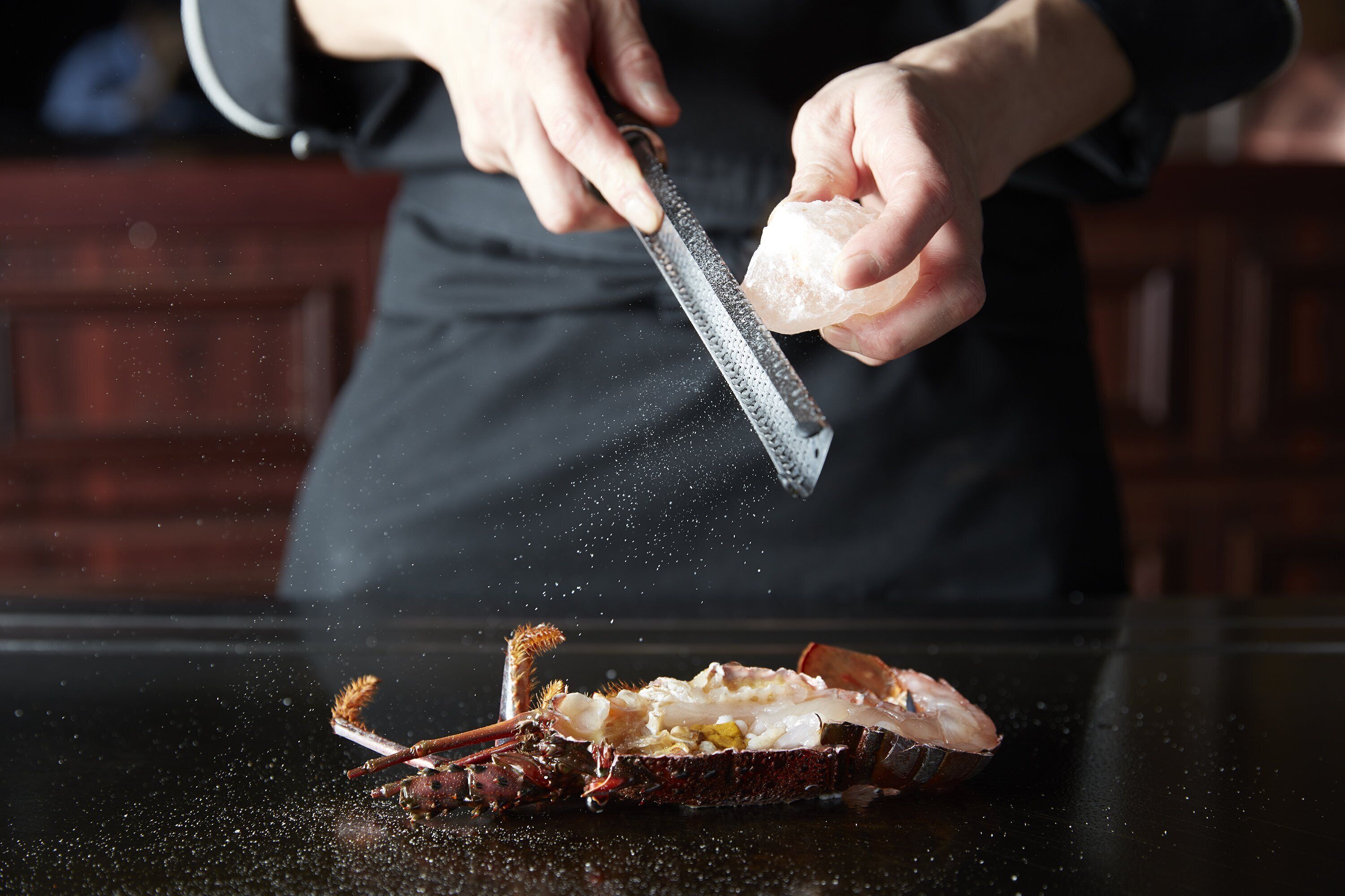 Una persona rallando una sustancia blanca, probablemente queso, sobre un langostino sobre una superficie negra.
