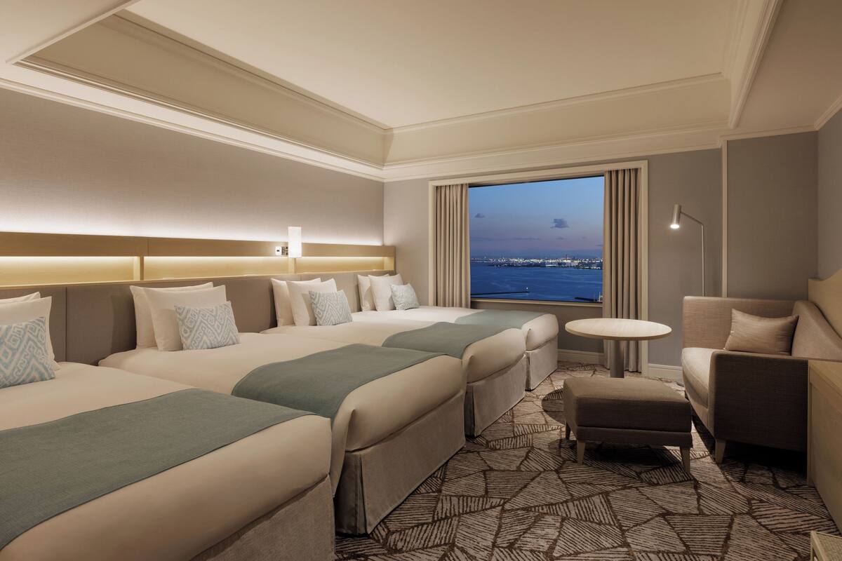 A hotel room with a large bed, two smaller beds, a sofa, a chair, a small table, and a view of the city at dusk.