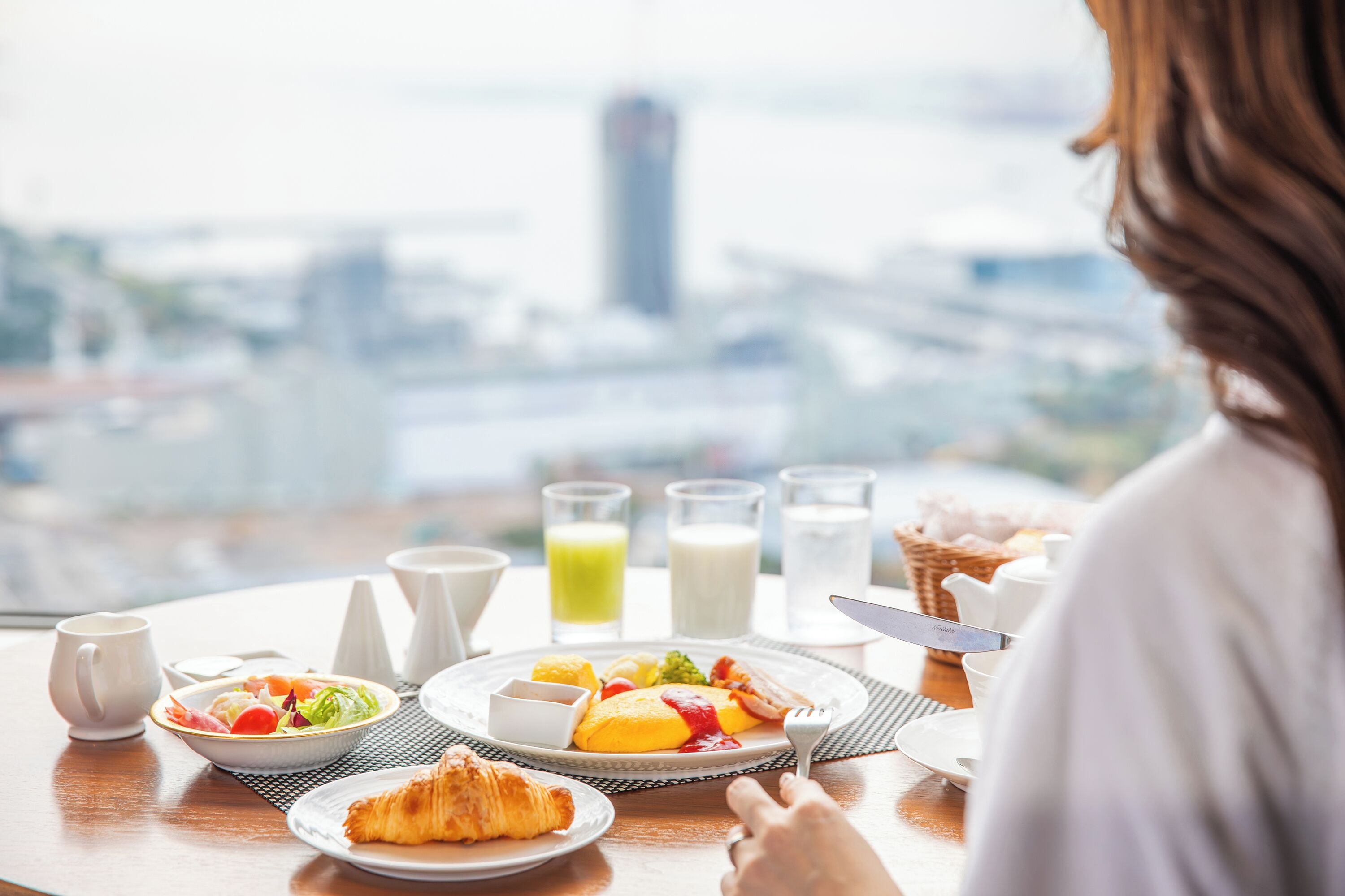 Ein Frühstückstisch mit Croissant, Obst und Getränken mit Blick auf eine Stadtlandschaft.